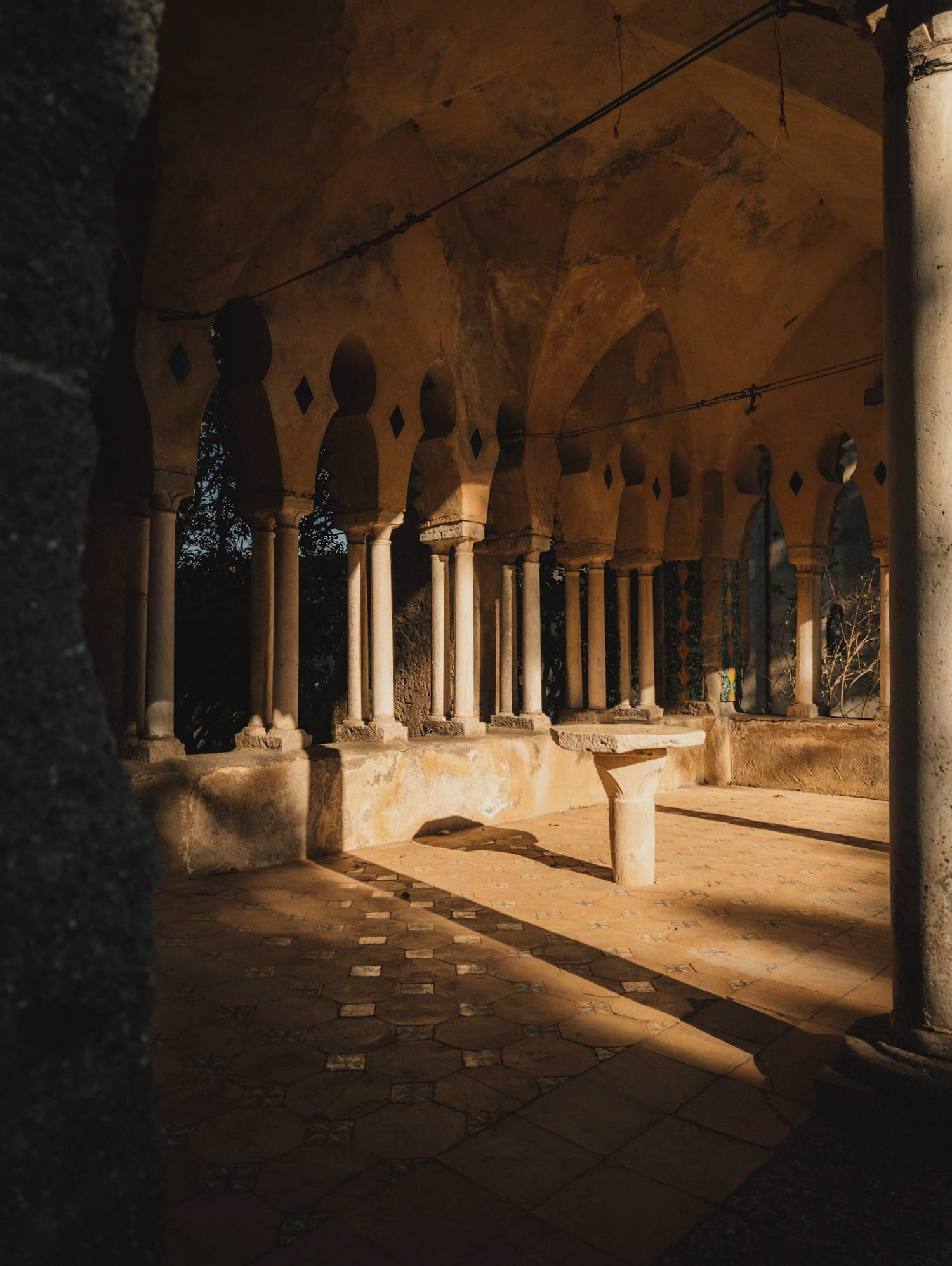 Sunlight falling through the arches of an old stone cloister onto a quiet colonnade.