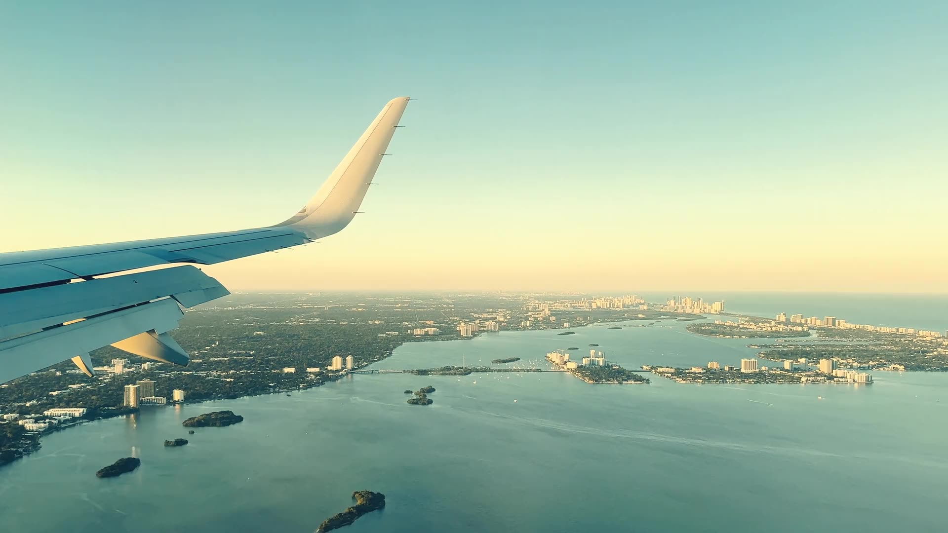 An aircraft in flight over turquoise water, the light going gold along the coast below.